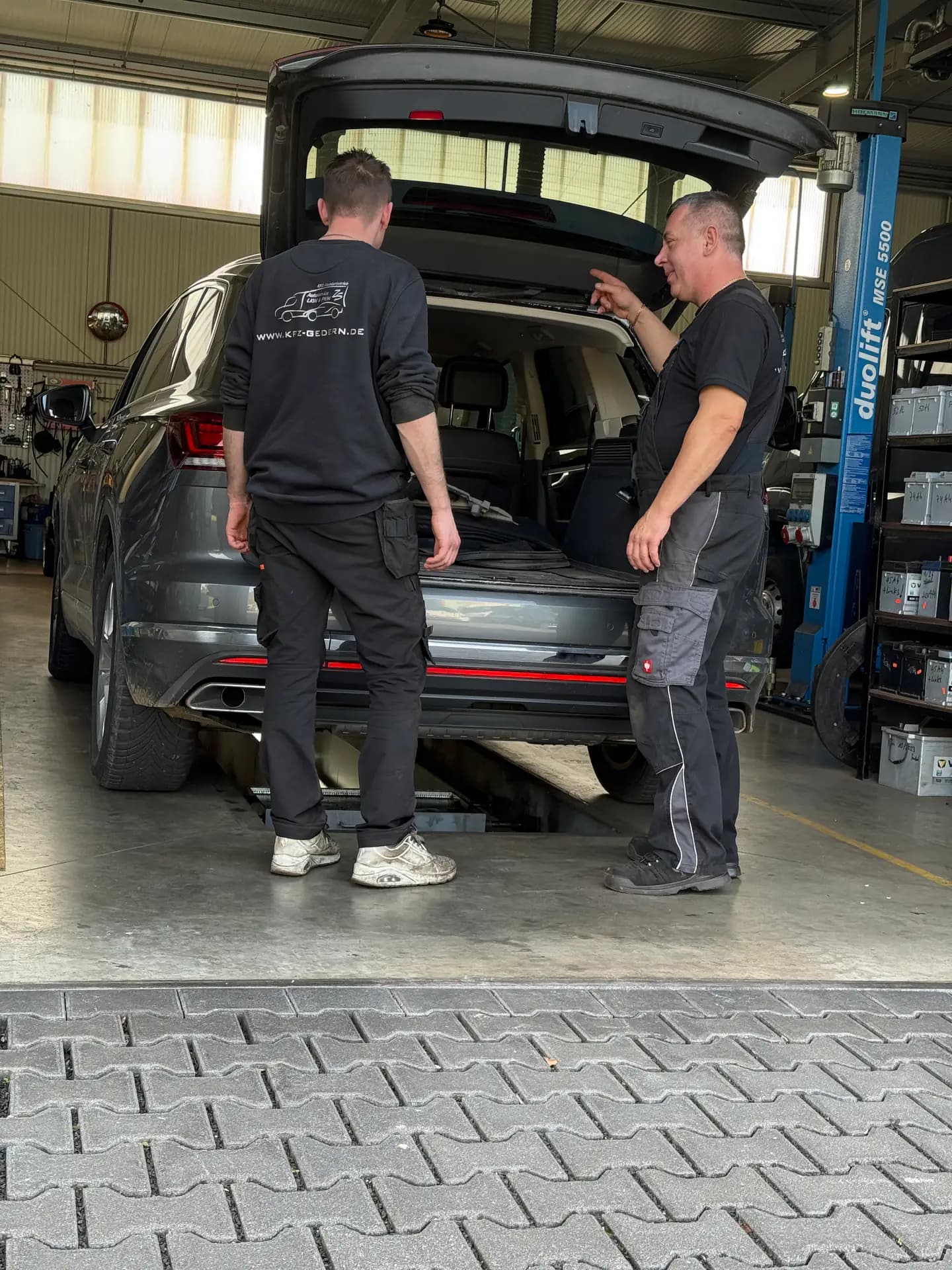 Meister erklärt einem jungen Kollegen etwas am offenen Kofferraum eines Autos auf der Hebebühne
